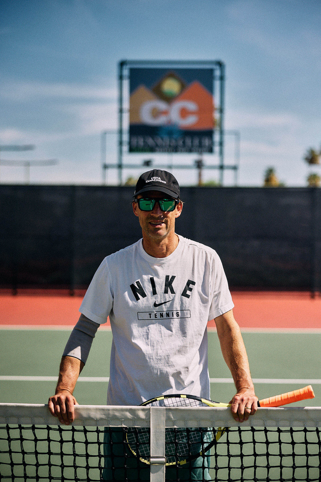 Coach Nathan Chesbro at the net in a Nike Tennis shirt and mirrored sunglasses, Cathedral Canyon club sign behind him.